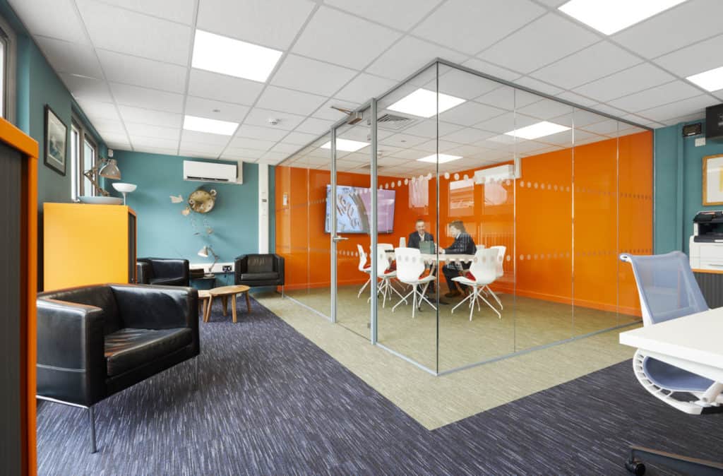 Inside a modular office. Meeting room with glass walls, a table and chairs and TV inside with two people sat at the table. Outside the meeting room there are 3 black leather arm chairs and orange cabinets. 