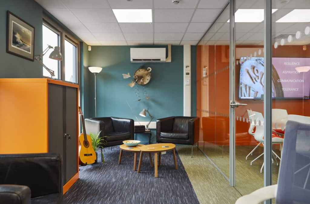 Inside modular school building. An office space, 2 black arm chairs around a small coffee table. Guitar next to an orange cabinet.
