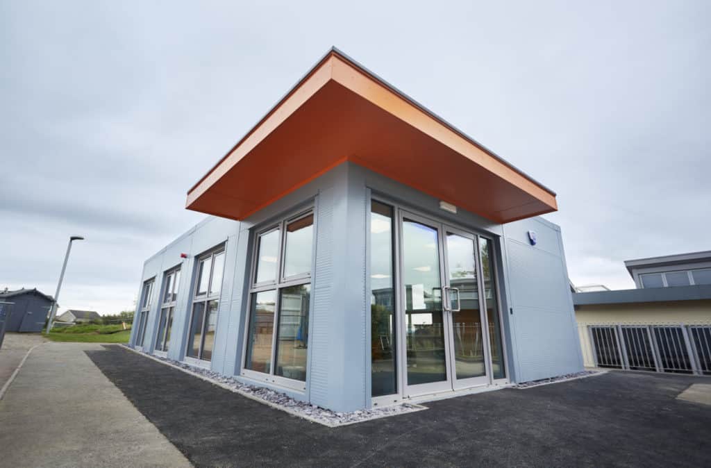 Modular eduction unit built to create more office space for staff. Orange canopy over a white box unit with several windows. Next to basket ball court. 
