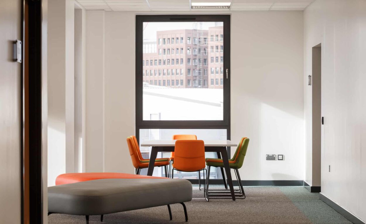 Inside modular classroom building at Leeds City College. 8 orange and green chairs surrounding a rectangular table, with two sofa stools.