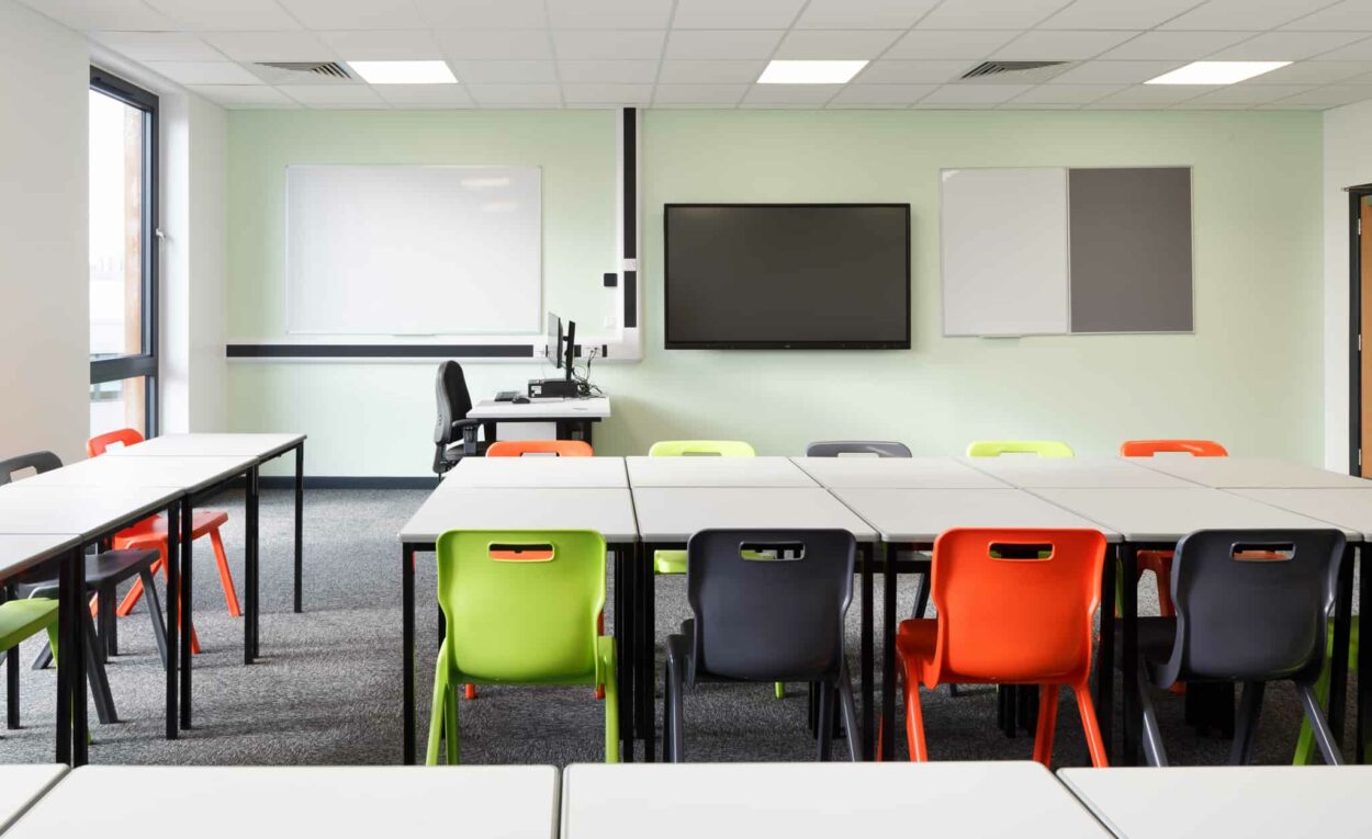 Inside a modular classroom. Orange, green and black chairs surround grey desks. Whiteboard and screen on the wall.