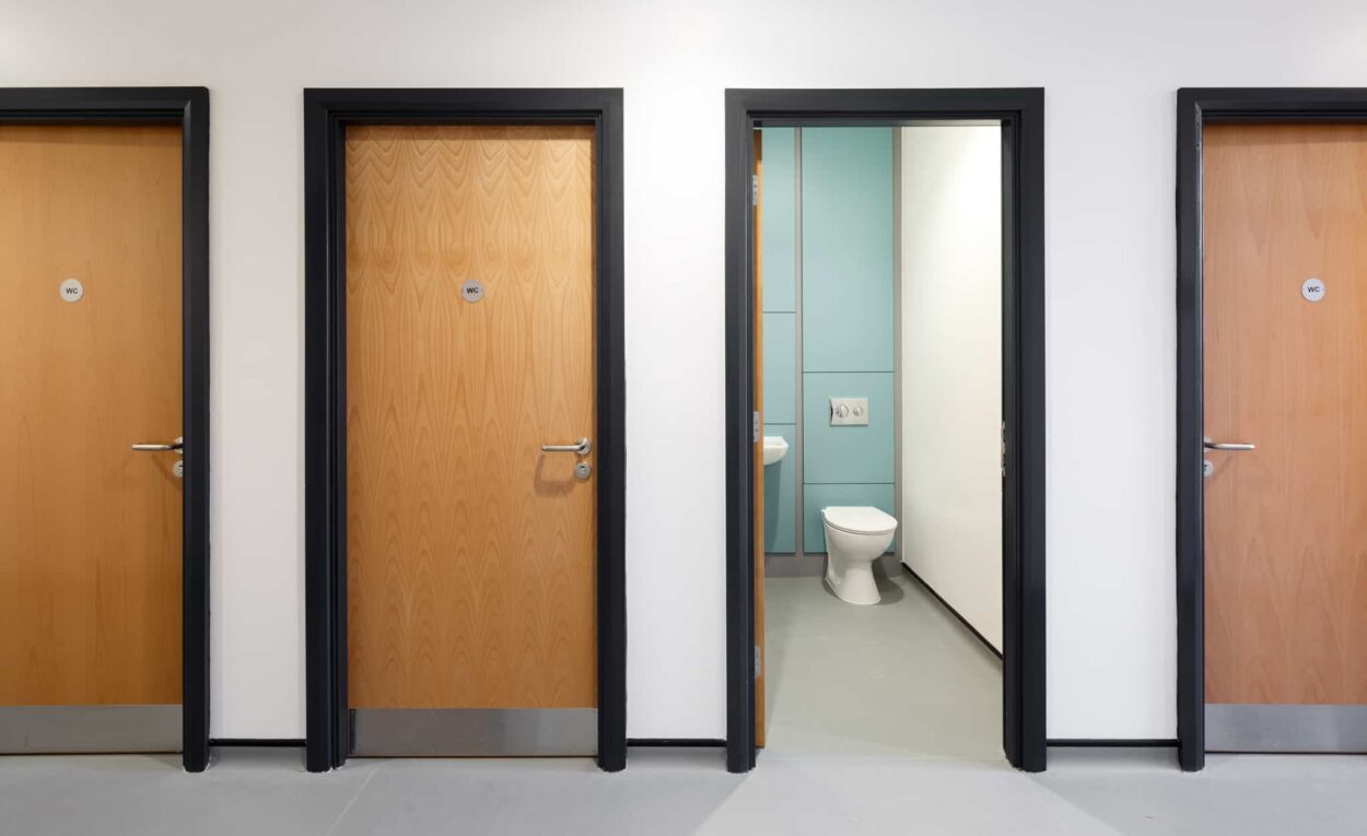Inside the modular school building where the toilets are. 4 individual toilet rooms, one door open with toilet showing, 3 doors closed.