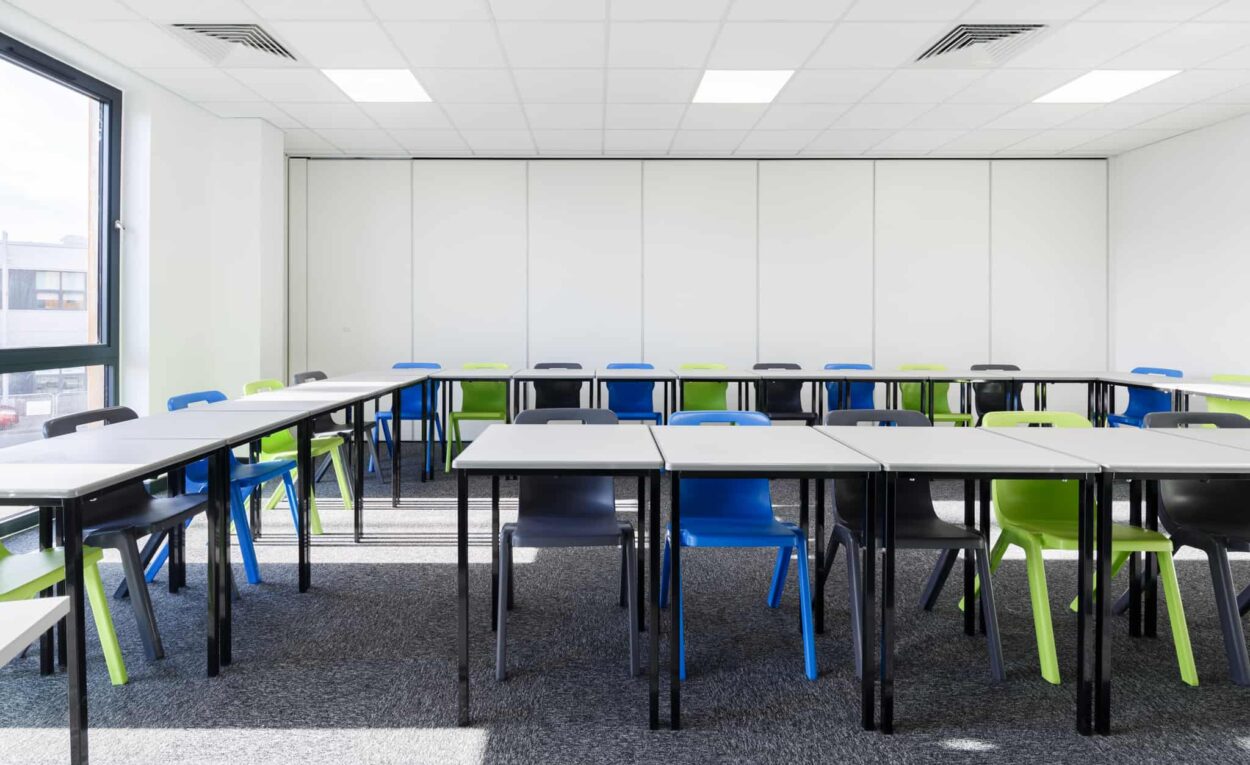 Modular classroom with white walls and several desks surrounded by black, green or blue chairs.